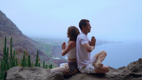 On-a-rock-at-the-mountain's-crest,-a-man-and-woman-meditate-and-practice-yoga-back-to-back,-gazing-out-at-the-ocean's-expanse